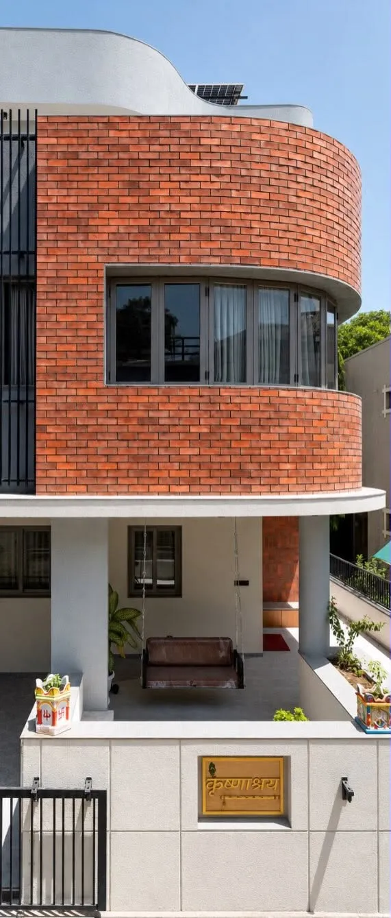 Close view of the Krishnashray facade: a curved exposed-brick drum wrapping a corner window, over a white-rendered porch with a traditional wooden swing and a brass nameplate.