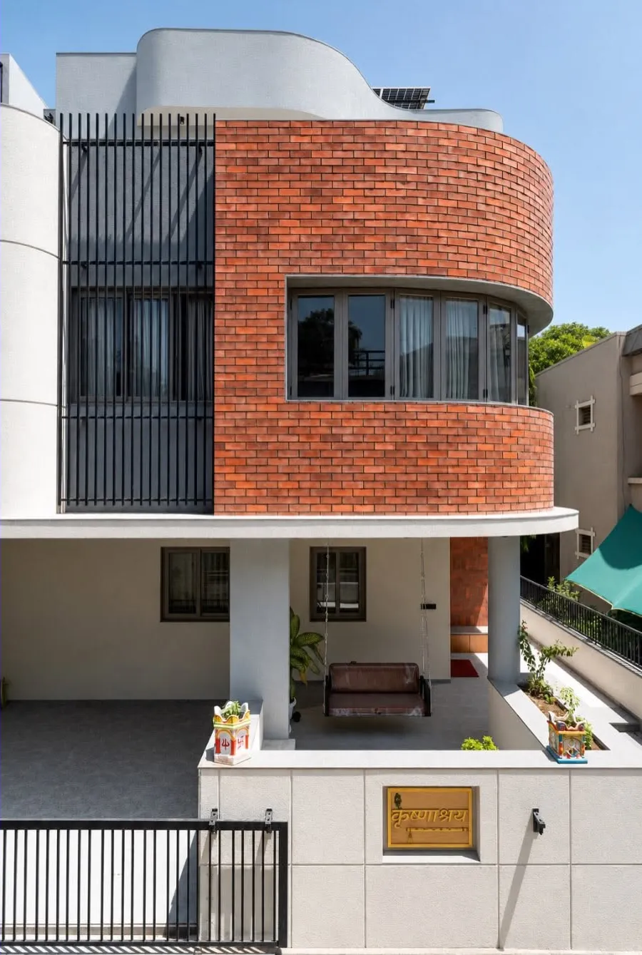 Full frontal elevation of Krishnashray showing the brick drum set against white rendered walls and a tall run of black vertical steel louvres.