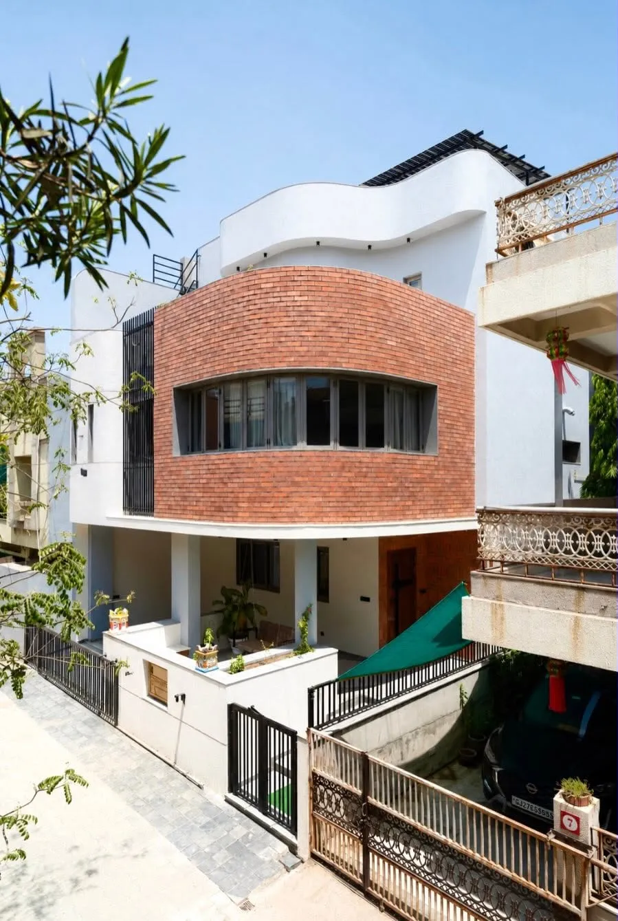 Krishnashray seen from the street between neighbouring houses, the warm brick volume reading against the white curved parapet and the sky.